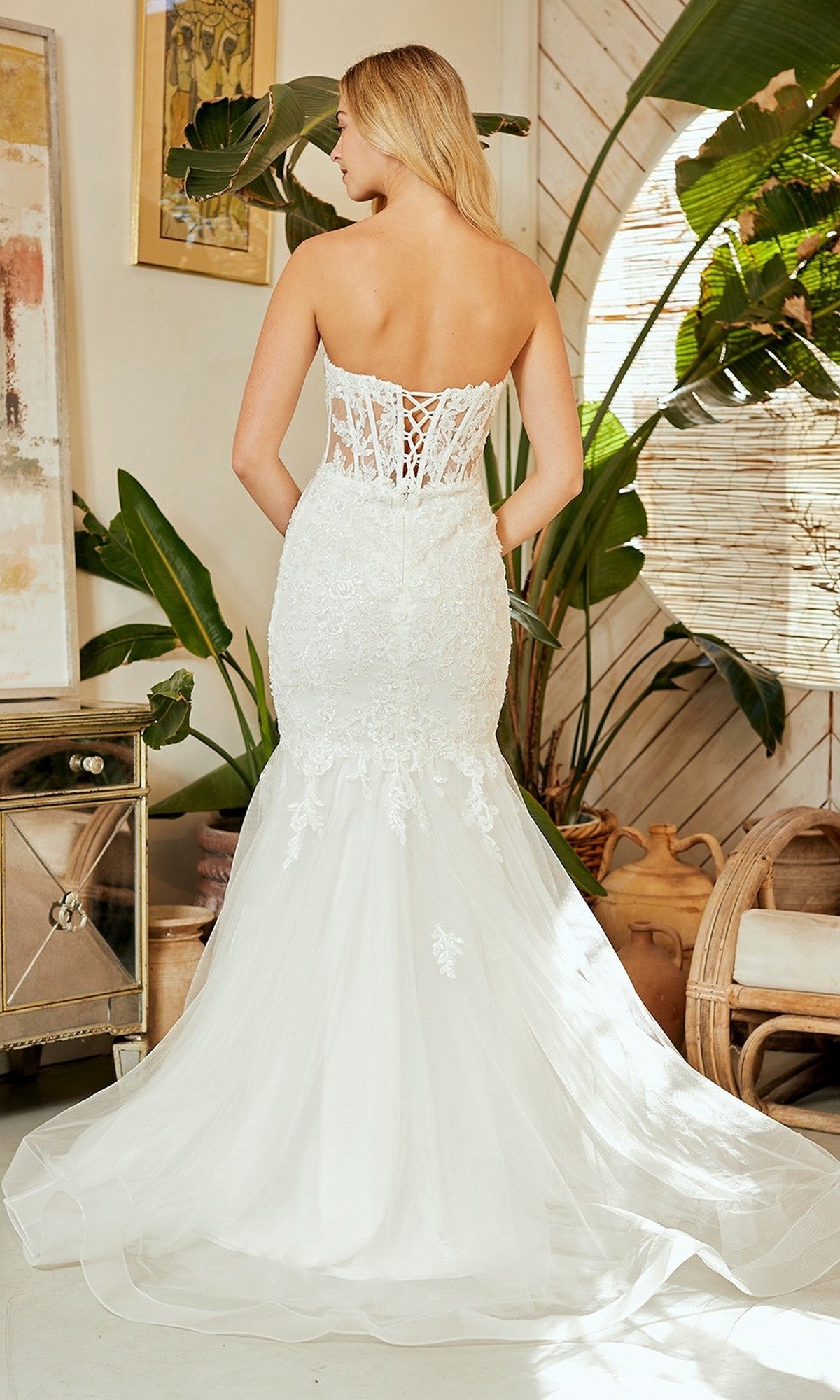 A woman in the Amelia Couture 7090 long prom dress stands indoors, facing away, surrounded by plants and neutral-toned decor. The dress features a strapless lace mermaid silhouette with an illusion corset bodice.